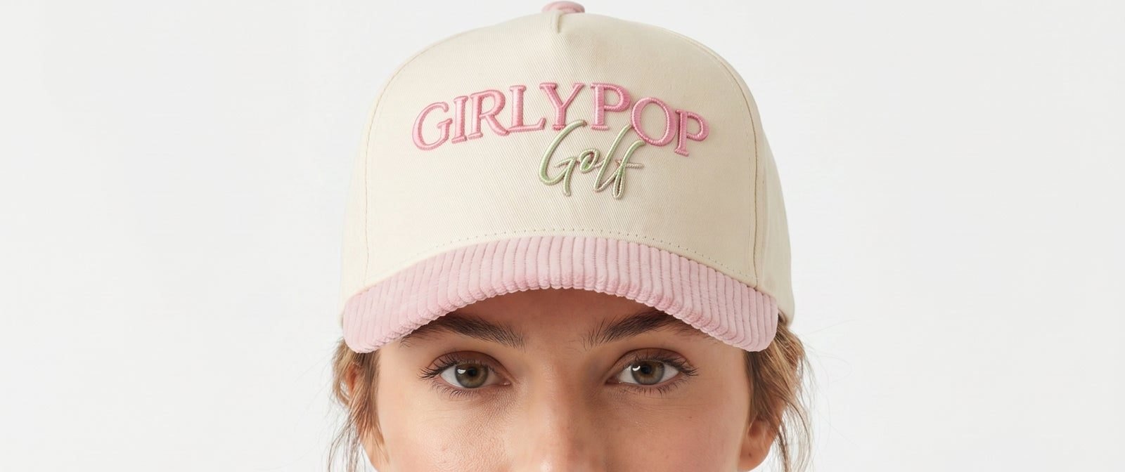 A woman looking straight at the camera under a cream Girly Pop Golf cap with a pink corduroy brim.