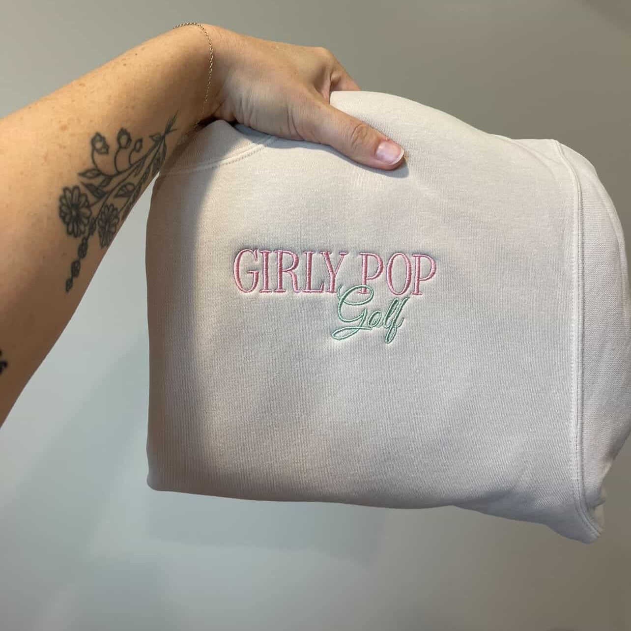 A hand holding a folded cream Girly Pop Golf sweatshirt with the embroidered logo showing.